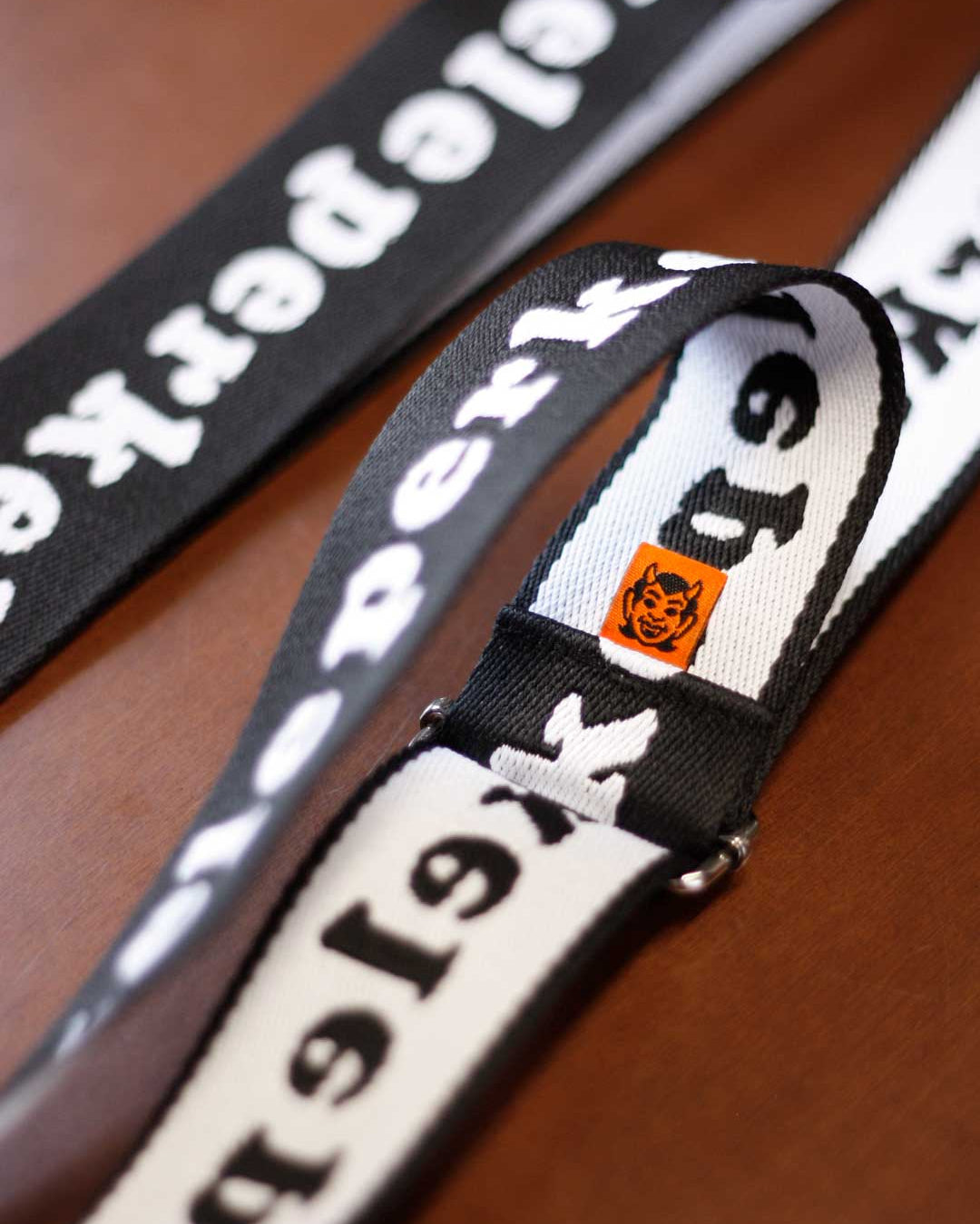 Close-up of a bag strap lanyard with perkele text and orange devil logo on a wooden surface
