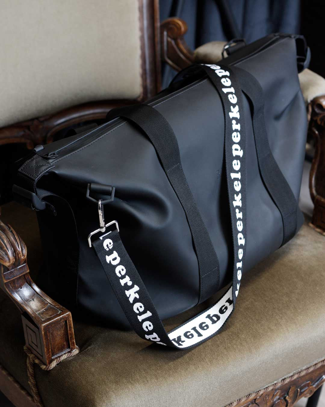 Black duffel bag with a visible perkele by kaamos brand strap on an ornate chair.