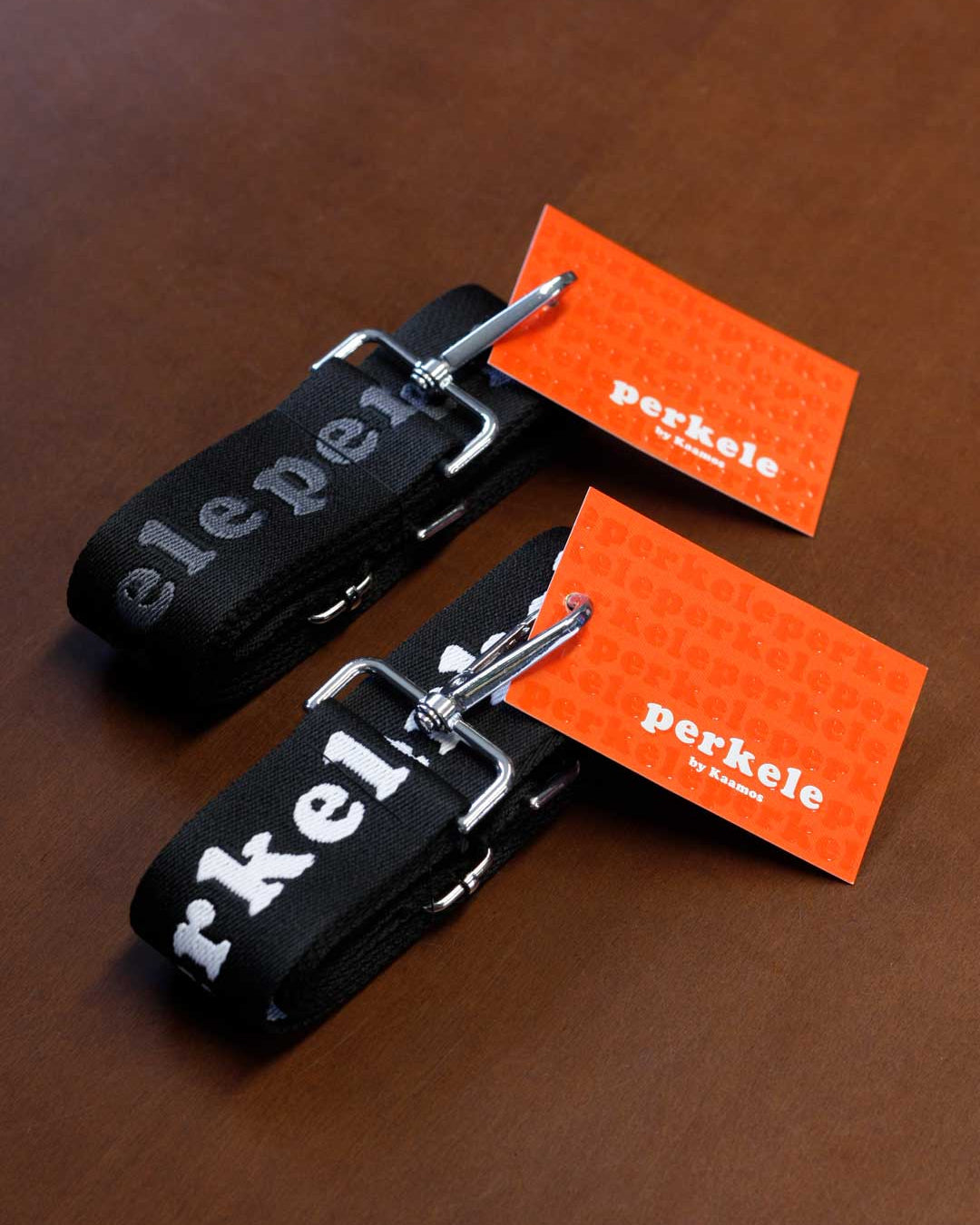 Two black keychains with silver clips on a brown surface, each accompanied by an orange card displaying 'perkele'.