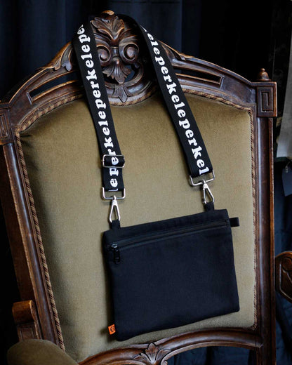 Black bag with interchangeable straps featuring 'perkele' text on a decorative chair.