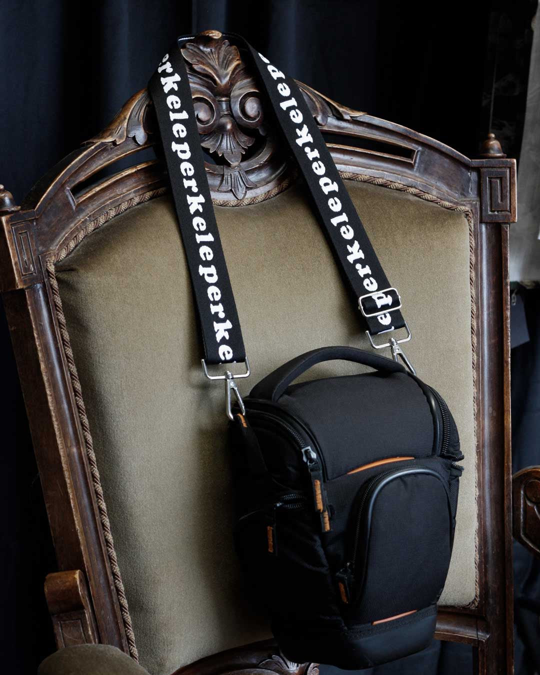 Black camera bag with 'perkeleperkele' strap on a decorative chair.
