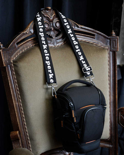 Black camera bag with 'perkeleperkele' strap on a decorative chair.